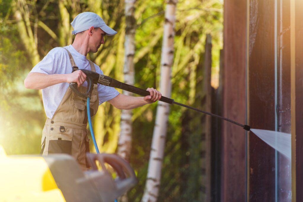 A person operates a pressure washer to clean the exterior of a house, spraying water on the walls and windows.