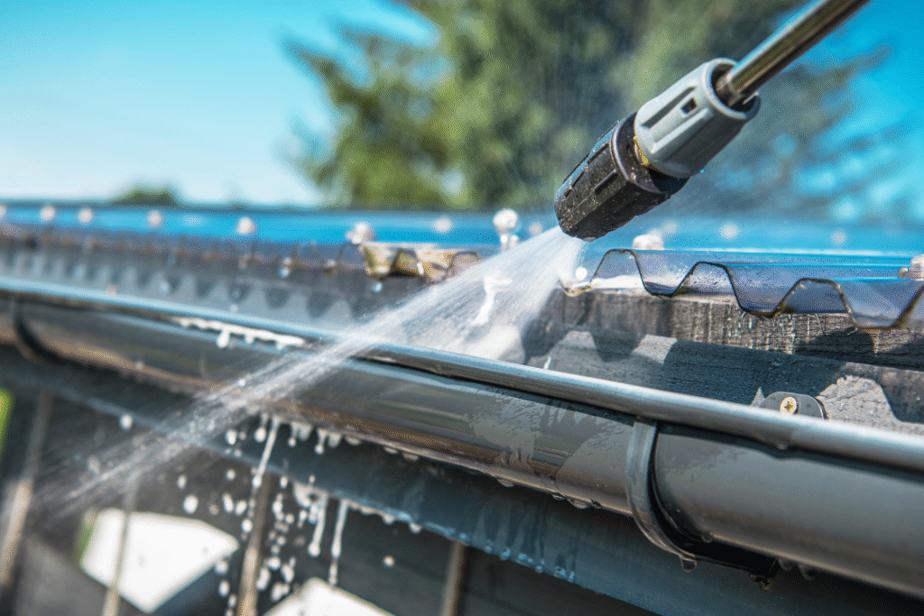 Close-up image of a pressure washer cleaning a metal gutter on a sunny day. Water sprays forcefully, creating a bright and refreshed appearance.
